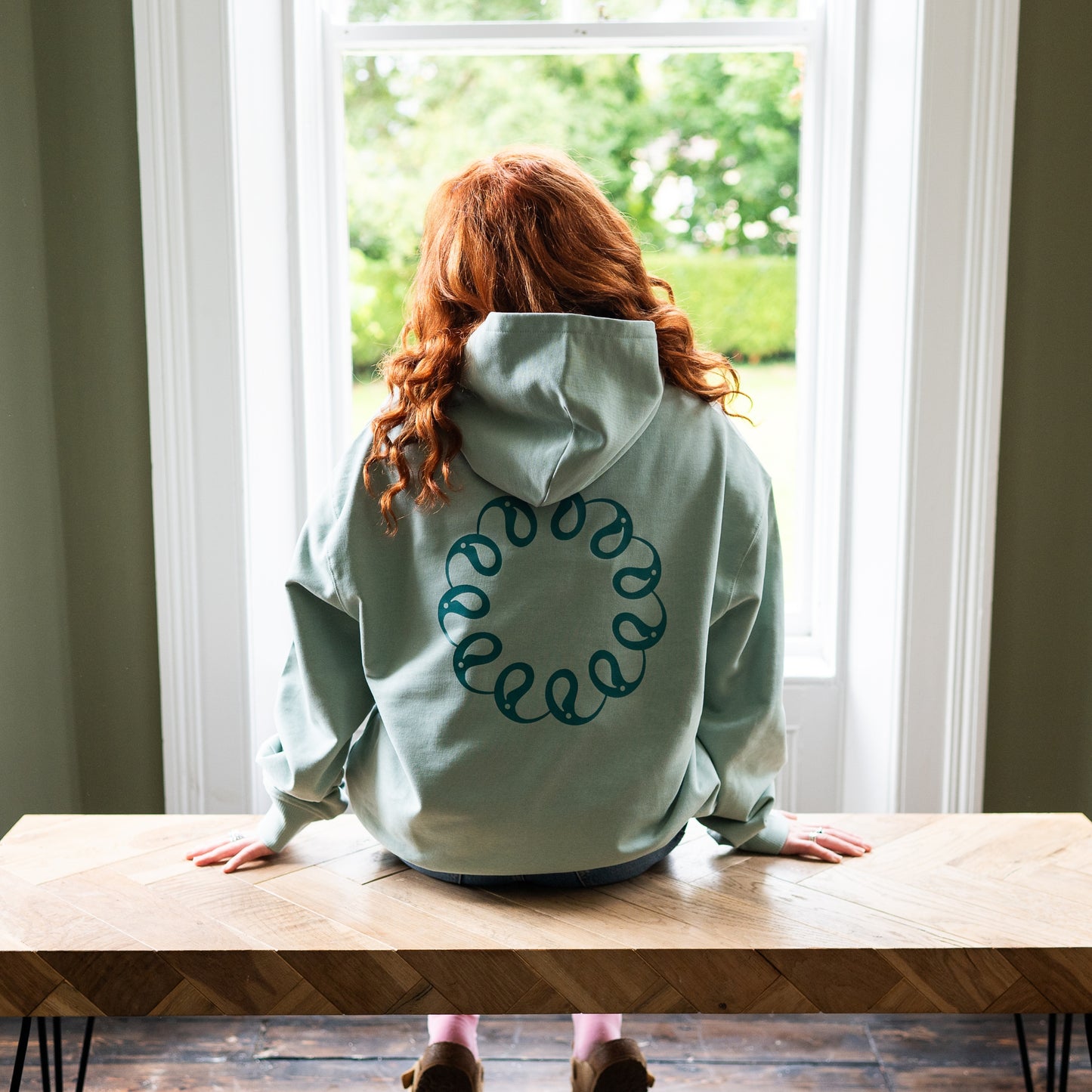 Girl sitting at window wearing Every Little Thread Hoodie in Aloe Colour. Back View of logo