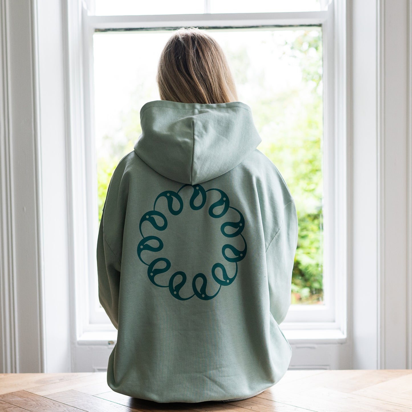 Girl sitting at window wearing Every Little Thread Hoodie in Aloe Colour. Back View of logo