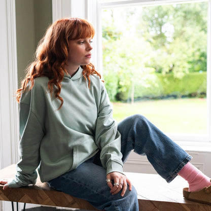 Girl sitting by a window wearing Every Little Thread hoodie in Aloe Colour