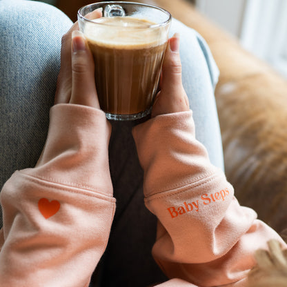 Person holding a glass of coffee with a wearing Baby Steps Sweatshirt in Peach colour by Every Little Thread