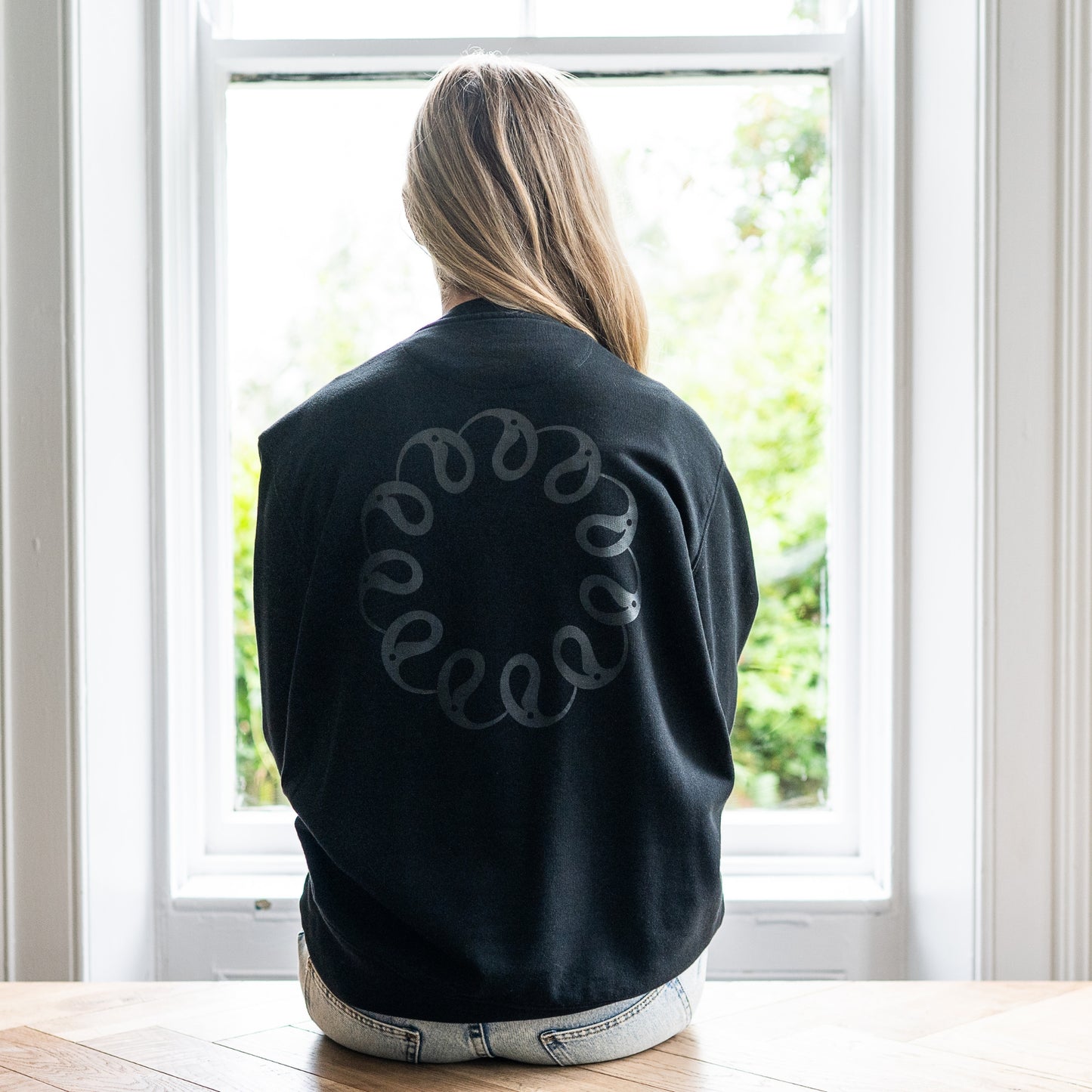 Girl sitting at window wearing Every Little Thread Sweatshirt in Black Colour. Back View of logo