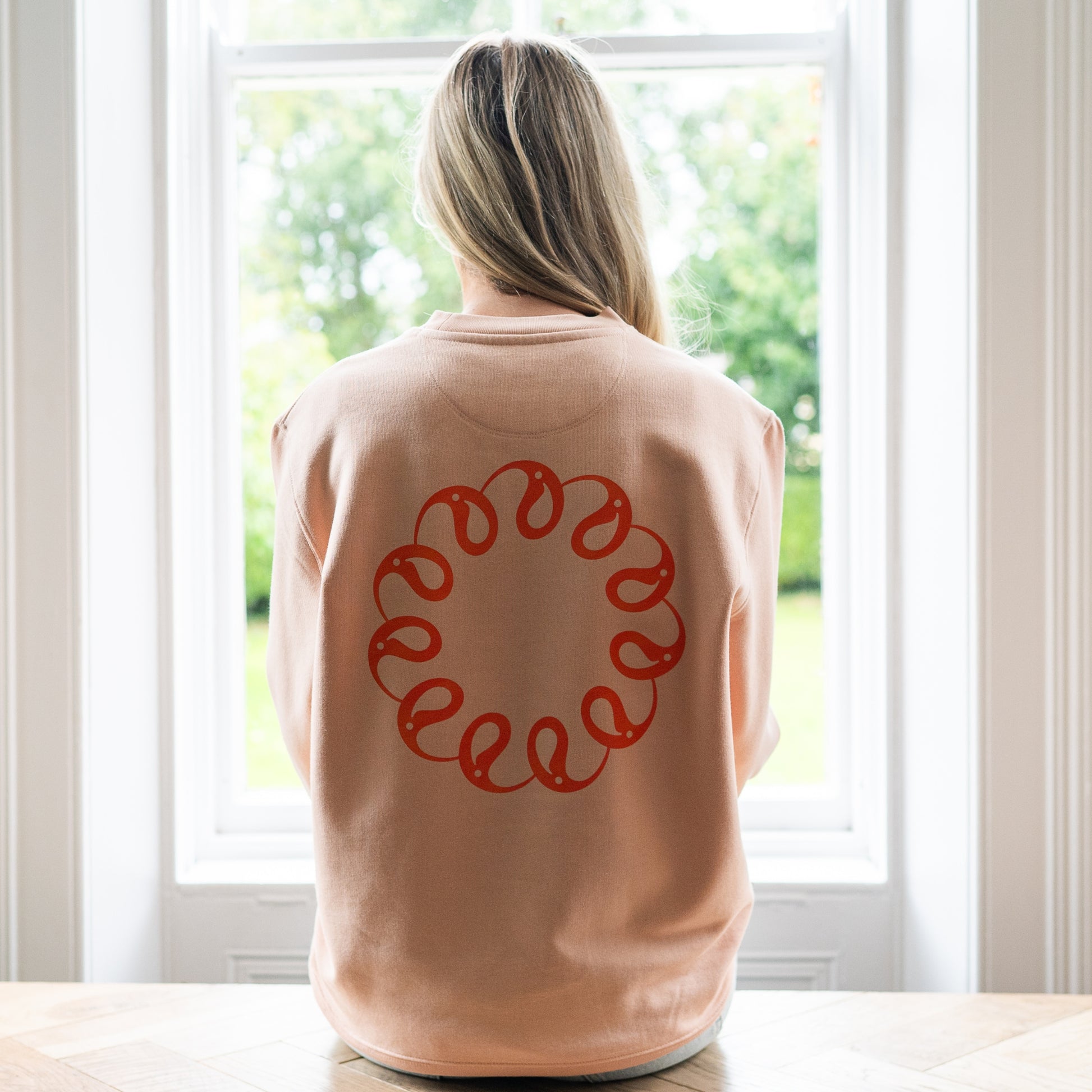Girl sitting at window wearing Every Little Thread Sweatshirt in Peach Colour. Back View of logo