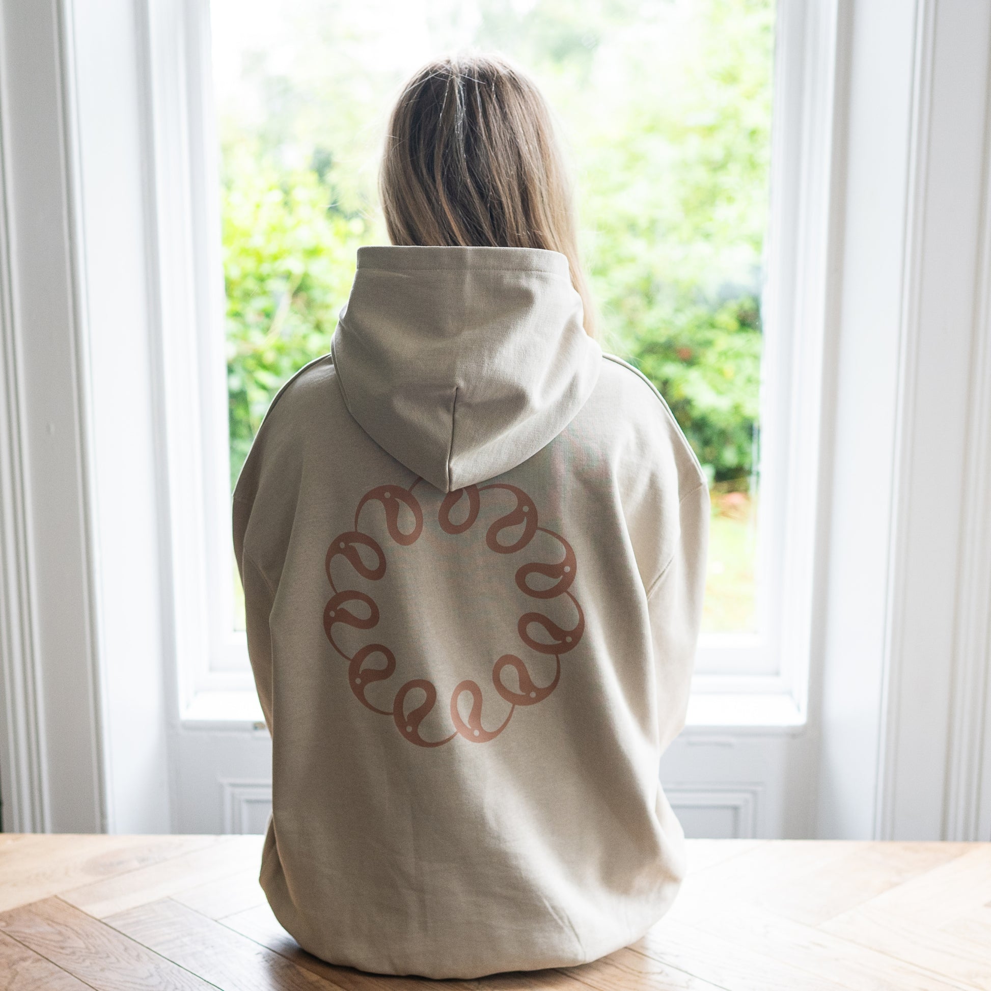Girl sitting at window wearing Every Little Thread Hoodie in Sand Colour. Back View of logo